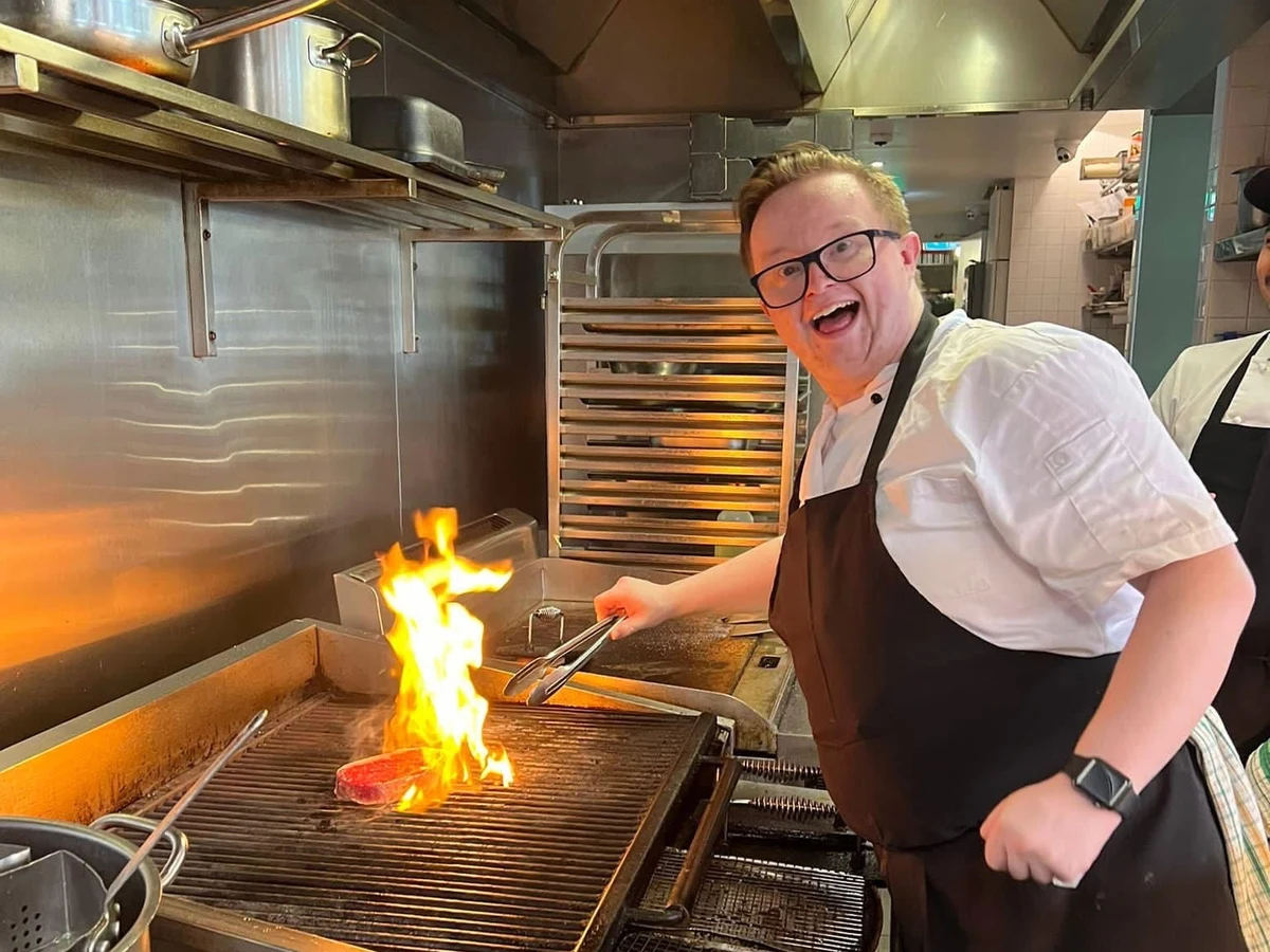 Tom in chef's whites and an apron, grilling at a commercial kitchen flat top.
